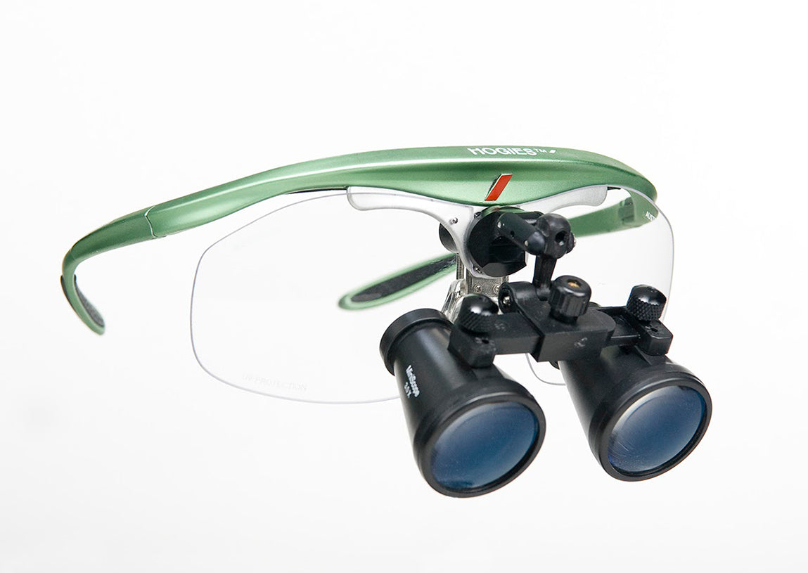 A pair of Hogies MiniScope loupes with green frame, black nose bridge, and clear glass lenses, along with additional accessories such as polycarbonate safety shields and an adjustment tool.