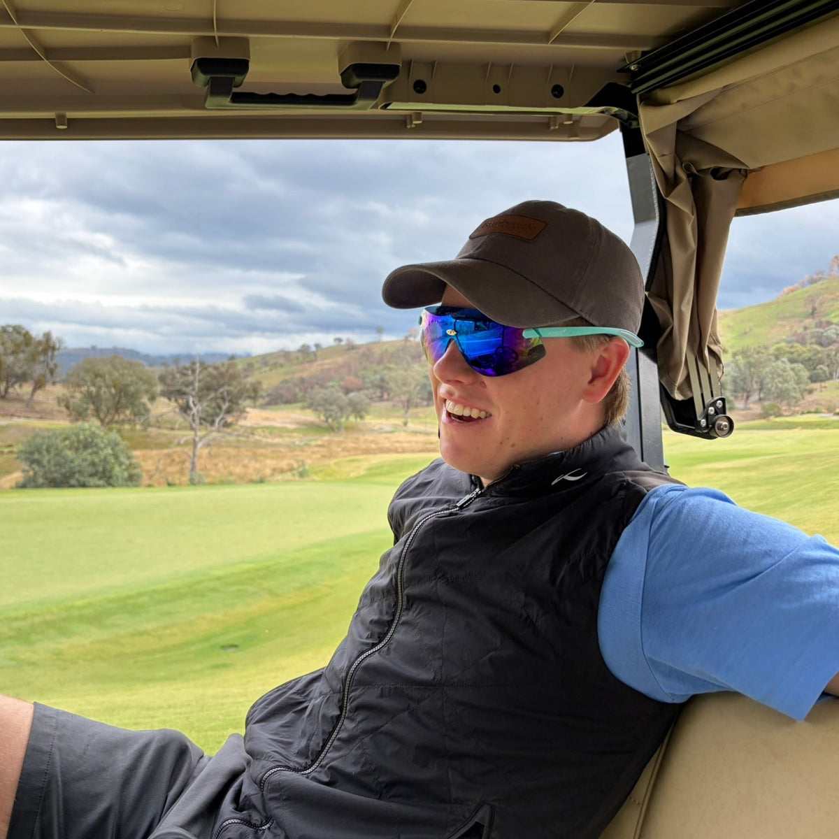 A man wearing sunglasses for golf sits inside a golf cart on a golf course.